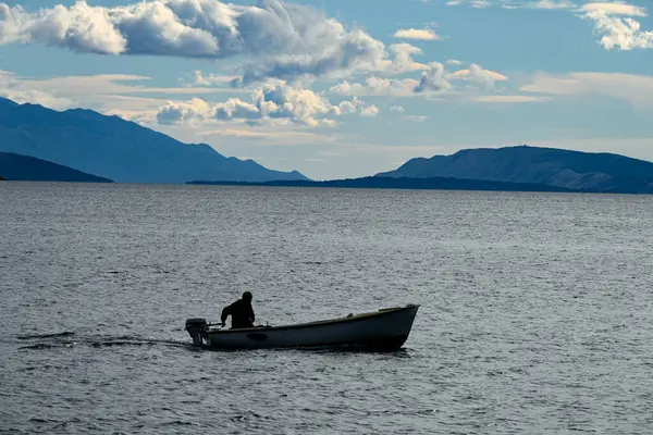 stock image a boat with a lone fisherman returning home after fishing on a calm sea under the rays of the sun setting behind the mountains.