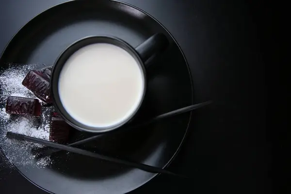 Organik sütle dolu büyük siyah bir seramik kap küçük siyah bir tabakta duruyor. Lezzetli meyveli marmelattan yapılan şekerlerle pudra şekeri serpiştirilmiş..