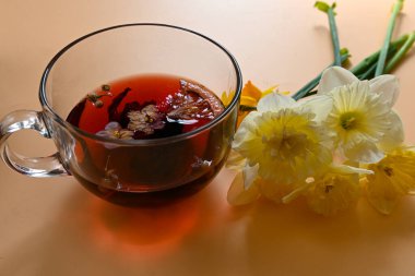 yakın plan cam şeffaf fincanda taze çay, papatya çiçekleri, portakal dilimleri ve amber çiçekleri, şeftali arkaplanda güzel sarı nergisler uzanır..