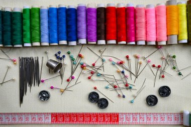 Bir terzinin işi için gerekli malzemelerin yakın çekimi: çok renkli iplikler, farklı boyutlarda iğneler, renkli iğneler, metal düğmeler, bir yüksük ve bir parça tebeşir ile kırmızı bant ölçüsü.