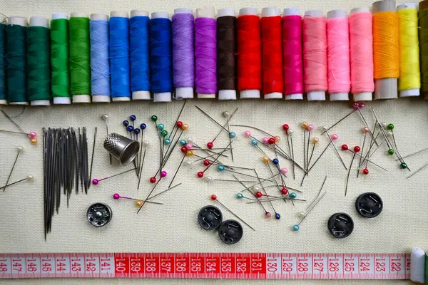 Bir terzinin işi için gerekli malzemelerin yakın çekimi: çok renkli iplikler, farklı boyutlarda iğneler, renkli iğneler, metal düğmeler, bir yüksük ve bir parça tebeşir ile kırmızı bant ölçüsü.