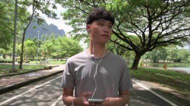 Asyalı yakışıklı çocuk yaz boyunca akıllı telefondan kulaklıkla müzik dinlerken halk parkında yürümekten zevk alır. Sağlıklı ve sağlıklı. Genç bir hayat tarzı. Açık hava aktivitesi.