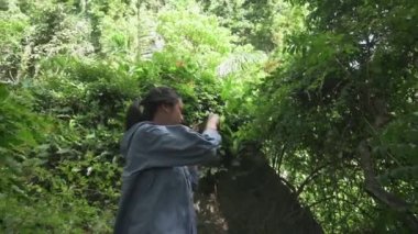Yaz boyunca yeşil tropikal yağmur ormanlarında cep telefonuyla video kaydeden sevimli Asyalı genç kız. Açık hava takibi. Boş zaman aktiviteleri. Genç yaşam tarzı. UHD.4K.