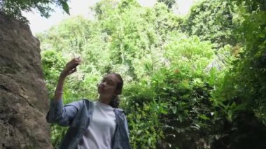 Asyalı sevimli genç kız tropikal yağmur ormanlarında yürüyüş yaparken cep telefonuyla selfie çekmeyi seviyor. Açık hava takibi. Boş zaman aktiviteleri. Genç yaşam tarzı. UHD. 4K.