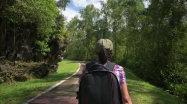 Asyalı serbest çalışan fotoğrafçının kamera çantasıyla parkta yürüdüğünü gösteren bir kamera görüntüsü. Yeşil bitkiler ve mavi gökyüzüyle halka açık bir parkta yaz manzarası. Açık hava takibi. Yavaş çekim.