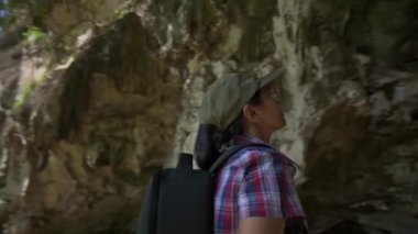 Dijital tabletli sıradan elbiseli jeolog kadın parkta kireçtaşı dağının oluşturduğu bir mağarayı keşfediyor. Arkanı dön ve geri çek. Hareketleri çek.. 