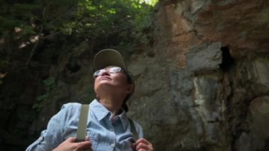 Orta yaşlı, sırt çantalı kadın kaşif yaz boyunca dik yamaçlarla çevrili dağda yürüyüş yaparken etrafına bakıyor. Yalnız macera. Açık hava takibi. Yörünge atışı.