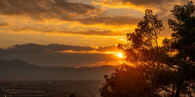 Dağların ve ağaçların üzerinde dramatik bulutlar ve ateşli gökyüzü ile altın gün batımı. Doğanın Güzelliğini ve Huzurunu Manzarada Yakalamak