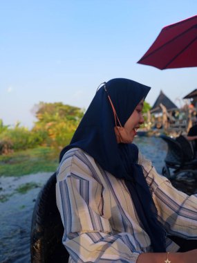 Kameranın önünde tesettürlü bir kadın, sakin ve rahat bir şekilde nehir kenarındaki bir sandalyede gülümsüyor ve bulanık bir arka planla tatilin tadını çıkarıyor.