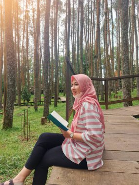Tesettürlü bir kadın, çam ormanında oturup kitap okuyor, yorucu bir işten tatilin tadını çıkarıyor, olumlu aktiviteler yapıyor, sessiz, rahat ve karanlık bir yerde bilgiyi arttırıyor.