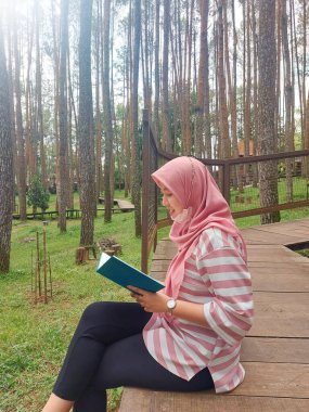 Tesettürlü bir kadın, çam ormanında oturup kitap okuyor, yorucu bir işten tatilin tadını çıkarıyor, olumlu aktiviteler yapıyor, sessiz, rahat ve karanlık bir yerde bilgiyi arttırıyor.