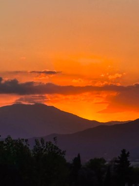 Dağ siluetleri ve bulutlar arasında parlayan güneş kremi ile turuncu gün batımı gökyüzü
