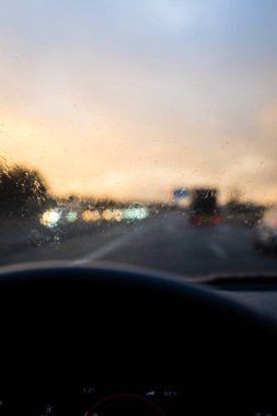 Yağmur damlalarıyla dolu bir arabanın ön camı. Alacakaranlık şoförünün bakış açısından yağmurlu bir yolda araba kullanırken.