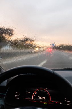 Yağmur damlalarıyla dolu bir arabanın ön camı. Alacakaranlık şoförünün bakış açısından yağmurlu bir yolda araba kullanırken.