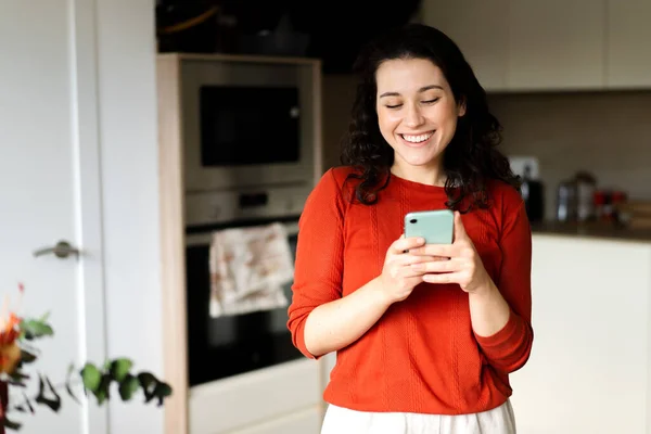 Młoda Szczęśliwa Kobieta Pomarańczowym Swetrze Używa Telefonu Komórkowego Stojącego Kuchni — Zdjęcie stockowe