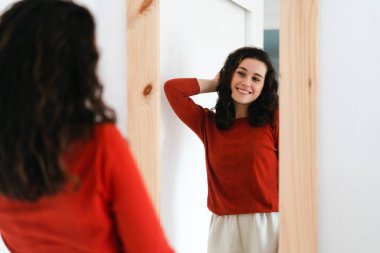  Kırmızı kazaklı, kendine güvenen genç ve güzel bir kadın aynaya bakıp kendisiyle gurur duyuyor.