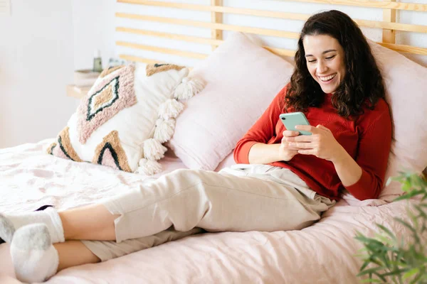 Yatakta Kırmızı Kazaklı Güzel Genç Bir Kadın Akıllı Telefonuna Bakıyor — Stok fotoğraf
