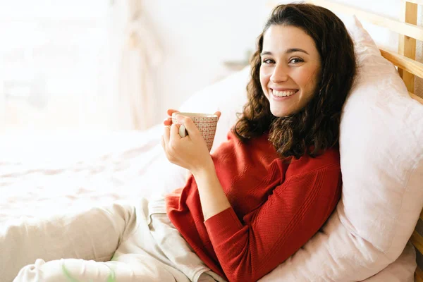 美しい若いです幸せな女性とともに赤いセーターリラックスしてベッドの上に横たわってコーヒーや紅茶 — ストック写真
