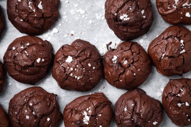 Ev yapımı browni kurabiyelerinin üst görüntüsü çatlamış üstü ve üzerine tuz parçaları serpiştirilmiş parşömen.