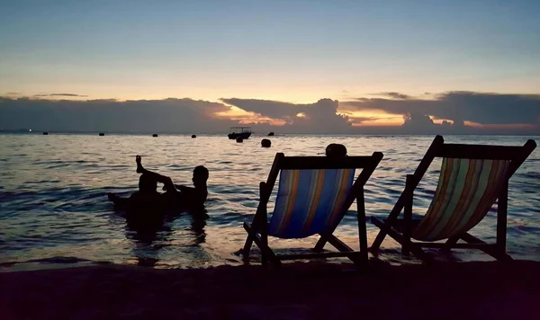 这张照片是两个人坐在沙滩上的椅子上 有两个人坐在沙滩上 水里有一条船 — 图库照片