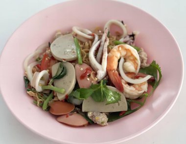 Salata ve karidesle dolu pembe bir kâse, beyaz bir masada karides, sebze ve erişte dolu bir tabak yemek..