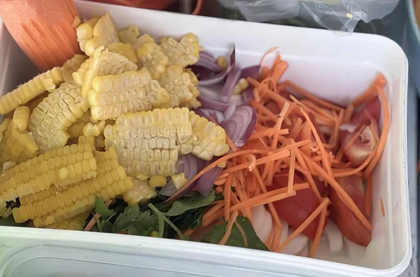 a photography of a container of vegetables and corn on a table, corn, carrots, onions, and other vegetables in a container.