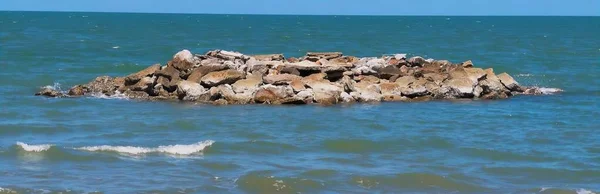a photography of a rock out in the ocean with a bird on top, lakeshore with rocks and waves in the ocean with a blue sky.