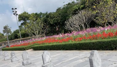 Bir sürü çiçek ve ağacın olduğu bir parkın fotoğrafı. Taş yolu olan bir parkta ağaçların ve çiçeklerin güneş saati..