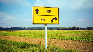 An image with a signpost pointing in two different directions in German. One direction is now, the other is never. clipart