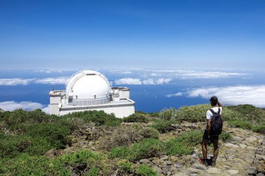 Hiker at the astronomical observatory of Roque de los Muchachos looking at Isaac Newton Telescope in a sunny day. Panoramic view with blue sky and clouds on the background.