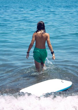 Uzun saçlı genç bir çocuk sörf tahtasıyla denize giriyor.