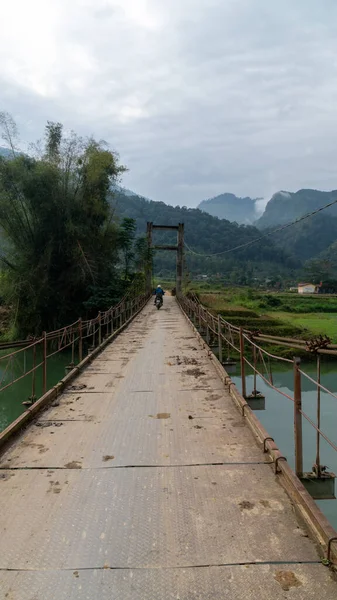 Ha Giang, Vietnam - 11 / 25 / 2019: Motosikletli adam nehirde küçük bir köprüden geçiyor. Turistler için popüler bir motosiklet döngüsü.