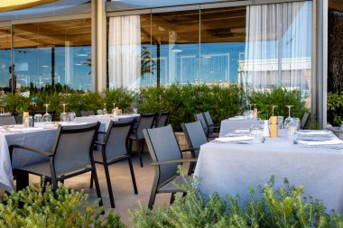 Modern bir tatil beldesi restoranının açık masaları. Bardaklar ve çatal bıçaklar yemek servisi için.