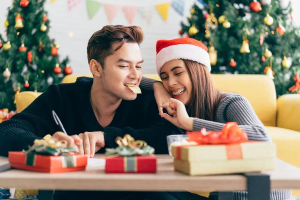 Asian man writing on a blank Christmas postcard with a pen while his girlfriend feeds him a potato chip. Couple sitting and writing Christmas card together at home during Christmas holiday.