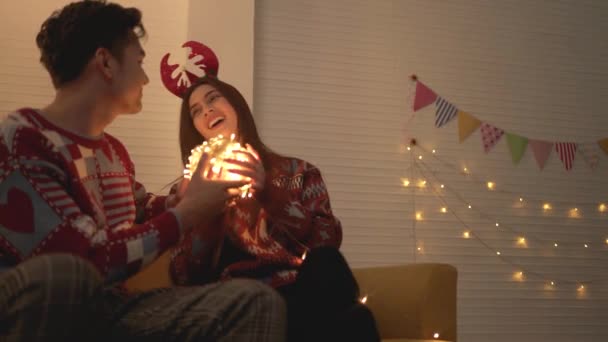 セーターを着たアジアのカップルは クリスマスの前夜に自宅のリビングルームのソファの上で一緒にLedライトボールを一緒に演奏しました クリスマスの夜に一緒に祝う — ストック動画
