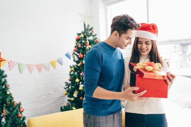 Noel Baba şapkası takan güzel, mutlu bir Asyalı kadın Noel hediyesiyle sevgilisine sürpriz yapıyor. Arka planda Noel ağacı var. Kopyalama alanı olan resim.