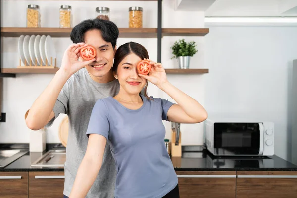 Healthy young Asian couple holding a fresh sliced tomato in their eye with a smile and happy emotion. An active lifestyle with healthy and vegetarian food advertisement. Image with copy space.