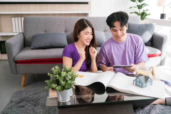 Young happy loving couple talking and using tablet to calculate the price of buying a house with a small wooden house model on a house drawing. New home concept.