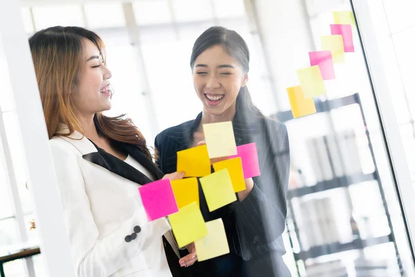 Group of successful Asian businesswomen teamwork. Brainstorm meeting with colorful sticky paper notes on the glass wall for new ideas. Using agile methodology for business in a tech start-up office.