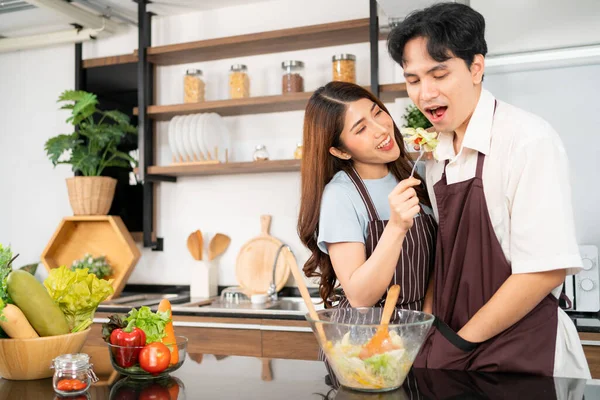 Happy Asian couple wearing aprons, a Woman feeding the vegetarian salad to a man for taste. Cooking a healthy salad with fresh vegetables in a glass bowl in the home kitchen.