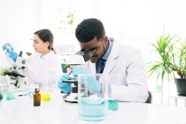 Genç Afrikalı siyahi bilim adamı araştırma yapmak için bir laboratuarda mikroskoba bakıyor. Öğrenciler biyokimyayı laboratuarda mikroskop araştırmalarında yeni ilaçlar bulmak için inceliyorlar..