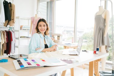 Kendine güvenen Asyalı bir terzinin portresi terzi atölyesinde giysiler üzerinde çalışıyor. Çekici bir bayan moda tasarımcısı gülümseyip arka planda elbise mankeni olan kameraya bakar..
