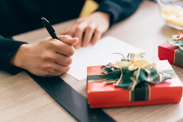Close up man\'s hand is writing on a blank Christmas postcard with a pen. Couple sitting and writing Christmas card together for sending with surprise gifts at home during Christmas holiday.