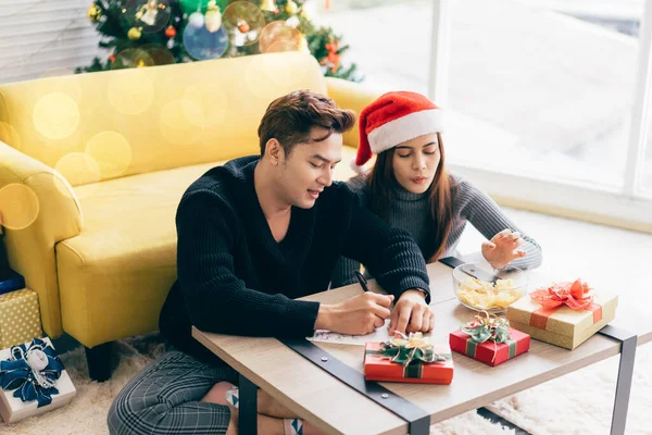 Asian man is writing on a blank Christmas postcard with a pen with his girlfriend. Couple sitting and writing Christmas card together for sending with surprise gifts at home during Christmas holiday.