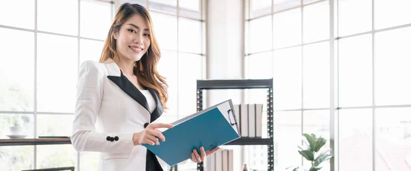 Confident and attractive Asian businesswoman wearing a professional look, holding a folder with a smile and looking at the camera in the office. Photo with copy space.