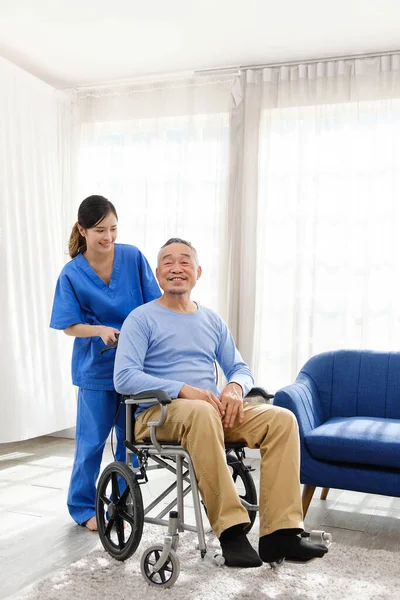 The caregiver therapist stands with an Asian senior sitting in a wheelchair with an empty sofa in the background. The nursing home facilitates a support group.