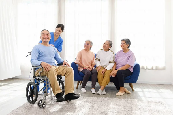 The caregiver therapist stands with an Asian senior sitting in a wheelchair with a group of senior women sitting on a sofa in the background. The nursing home facilitates a support group.