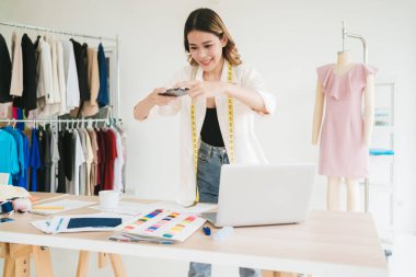 Asyalı kadın moda tasarımcısı ya da terzi masada duruyor, müşteriye göndermek için akıllı telefondan kumaşların ve örnek materyallerin fotoğraflarını çekiyor. Bir atölyede yeni koleksiyonlar tasarlayan terzi.