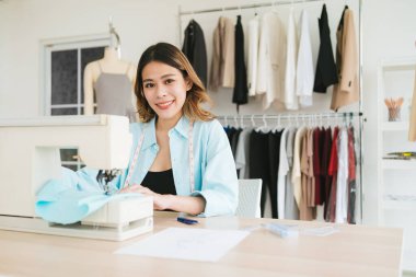 Kendine güvenen Asyalı bir terzinin portresi dikiş makinesiyle elbise dikiyor. Genç ve çekici bir bayan moda tasarımcısı gülümseyecek ve arka planda elbise askısı olan kameraya bakacak..