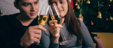 Mutlu Asyalı çift şampanya kızartıyorlar, akşamları koltukta oturuyorlar ve arka planda bir Noel ağacı ve ışıklar var. Noel arifesinde sevgili